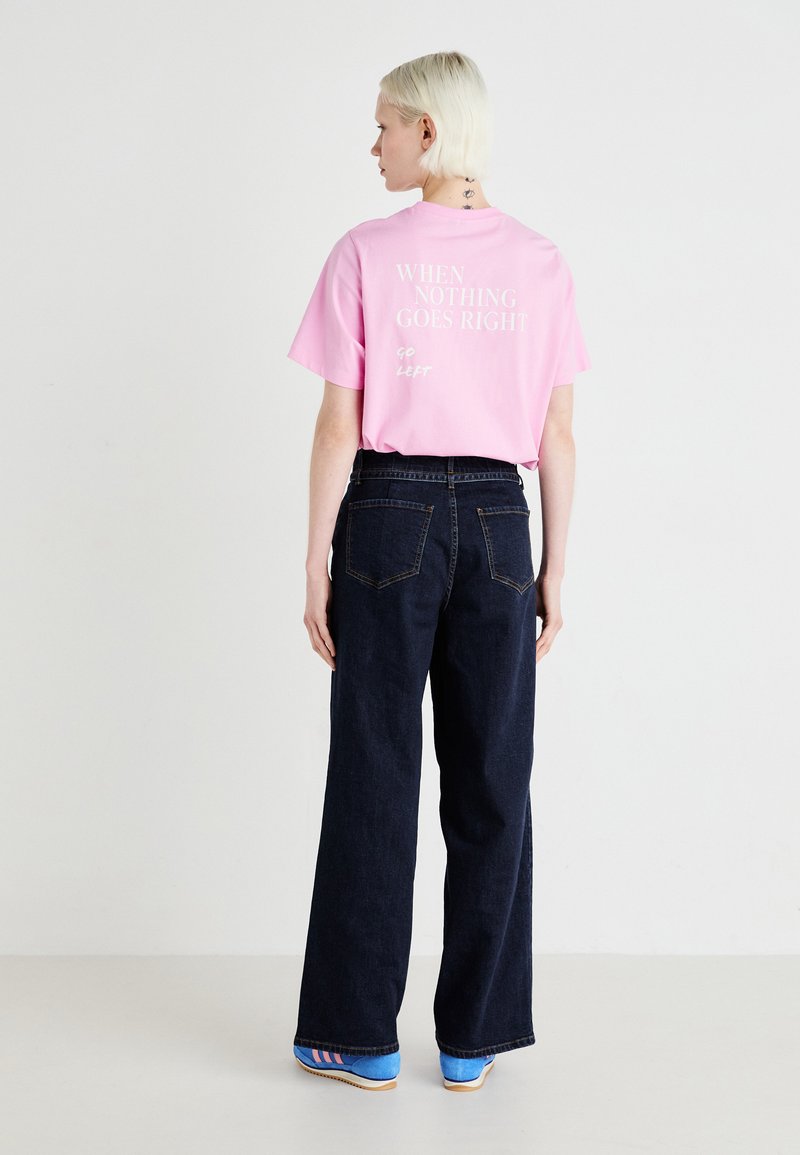 Roze katoenen t-shirt met witte tekst op de achterkant, gecombineerd met donkerblauwe wijde jeans en blauwe schoenen met roze strepen.