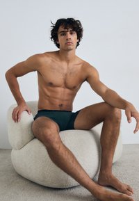 Modello maschile seduto su una sedia morbida e rotonda, indossando pantaloni corti attillati di colore verde scuro. Lo sfondo è bianco uniforme e il pavimento è strutturato.