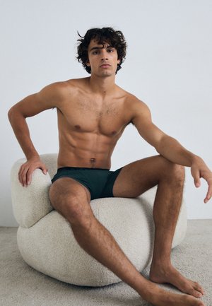 Modello maschile seduto su una sedia morbida e rotonda, indossando pantaloni corti attillati di colore verde scuro. Lo sfondo è bianco uniforme e il pavimento è strutturato.