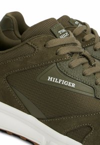 Zapatillas verde oliva que combinan materiales de gamuza y textil, con una media suela blanca y un destacado logo de la marca en el lateral.