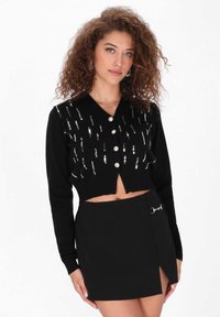 Schwarze, verkürzte Strickjacke mit silberner Paillettenverzierung, V-Ausschnitt und fünf Knöpfen. Kombiniert mit einem schwarzen Minirock mit einem seitlichen Schlitz und Hardware-Akzenten.