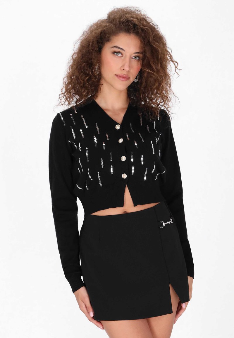 Schwarze, verkürzte Strickjacke mit silberner Paillettenverzierung, V-Ausschnitt und fünf Knöpfen. Kombiniert mit einem schwarzen Minirock mit einem seitlichen Schlitz und Hardware-Akzenten.