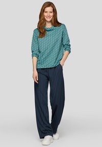 Junge Frau mit langen roten Haaren trägt ein türkisfarbenes Oberteil mit geometrischem Muster, marineblaue, gestreifte Culotte und weiße Turnschuhe, steht mit der Hand in der Tasche.