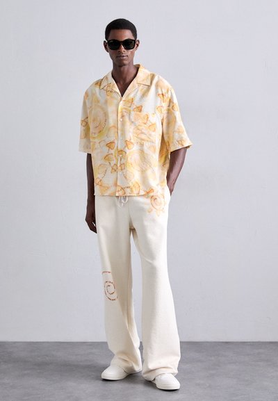 Homme portant des lunettes de soleil noires, une chemise à manches courtes jaune clair avec des motifs oranges, un pantalon blanc ample avec des accents oranges, et des baskets blanches.