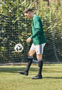 Groene lange mouwen sporttrui, witte shorts met zwarte strepen, zwarte voetbalschoenen en zwarte sokken. Bal in beweging op een grasveld.