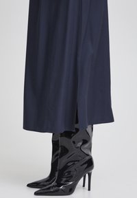 Bottes montantes en cuir verni noir à talons aiguilles avec des orteils pointus, associées à un vêtement noir bleu foncé longueur cheville présentant une fente sur le côté.