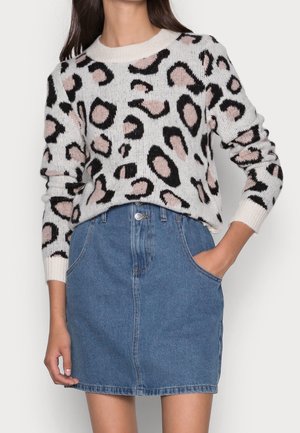 Femme portant un pull blanc avec un imprimé léopard rose, glissé dans une jupe en jean bleue à poches avant.
