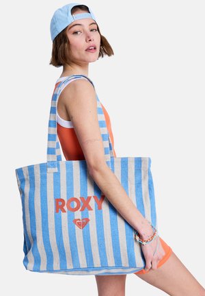 Joven mujer con gorra azul y conjunto naranja sosteniendo un bolso grande de rayas azules y beige con el logo "ROXY".