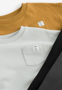 Drie crewneck sweaters in mosterdgeel, lichtgrijs en zwart. Kenmerken zijn ribgebreide kragen, textuurstof en kleine labels op de voorkant.