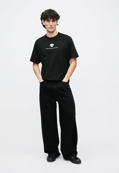 Μαύρο βαμβακερό T-shirt με γραφικό τύπωμα στο στήθος, σε συνδυασμό με φαρδιά μαύρη παντελόνα. Το ντύσιμο χαρακτηρίζεται από μια χαλαρή σχεδίαση.