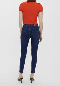 Top corto de manga corta de rayas rojas emparejado con jeans ajustados de color azul oscuro. Los jeans cuentan con dos bolsillos traseros y el modelo lleva sandalias de tacón verdes.