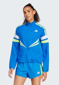 Veste de survêtement bleue avec des rayures vertes et blanches, présentant un col haut et une fermeture éclair à l'avant. Accompagnée de shorts bleus assortis, tous deux avec le logo Adidas.