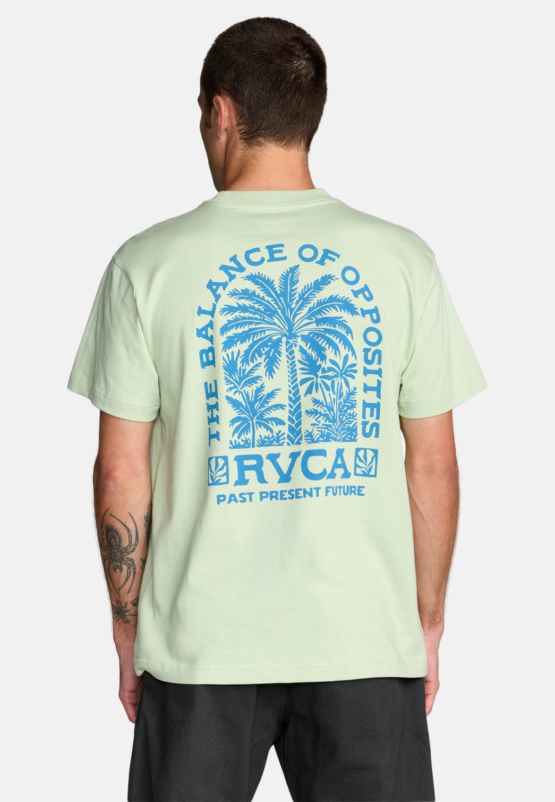 Mann trägt hellgrünes T-Shirt mit blauem Palmenmotiv und dem Text "The Balance of Opposites RVCA Past Present Future" auf der Rückseite.
