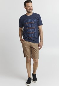 Marineblå T-skjorte med hvit og oransje tekst, kombinert med lysebrune shorts. Svarte joggesko fullfører det uformelle antrekket.
