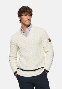 Jersey de punto trenzado en color crema con diseño de escote en pico, tejido texturizado, franja en color marino en la parte inferior y parche bordado en la manga izquierda.