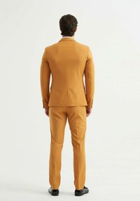 Costume pour homme de couleur moutarde vif. Veste cintrée avec revers crantés et pantalon slim-fit. Texture lisse et design minimaliste.