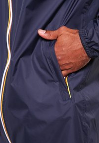 Laivastonsininen tuulitakki, jossa on vetoketjullinen tasku värikkäällä korostusreunuksella, sileä kangaspinta ja joustavat hihansuut varmistamassa napakan istuvuuden.