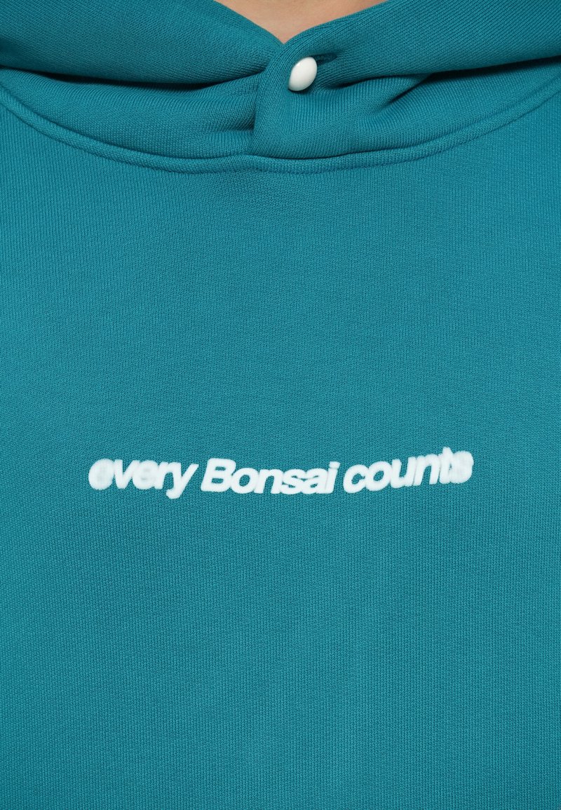 Türaqua Kapuzenpullover mit weicher Textur, auf dem die eingestickte Phrase "Jeder Bonsai zählt" in Weiß, mittig auf der Brust, platziert ist.