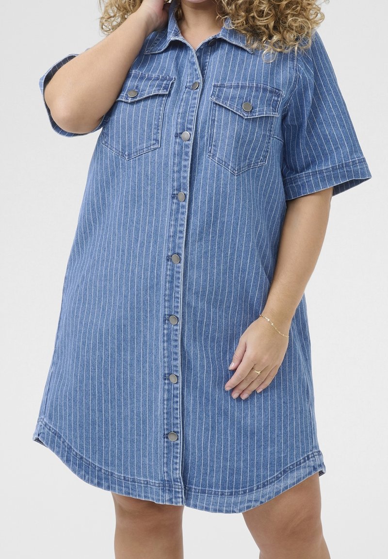 Femme portant une robe en denim bleue à manches courtes avec des fines rayures blanches et des boutons-pression métalliques sur le devant, debout devant un fond uni.