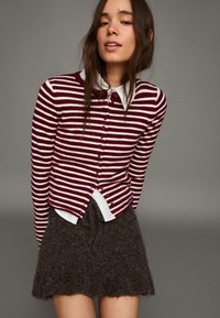 Bordeauxrood-wit gestreepte cardigan met knoopsluiting, gecombineerd met een getextureerde donkergrijze wollen rok en een wit overhemd met kraag eronder.