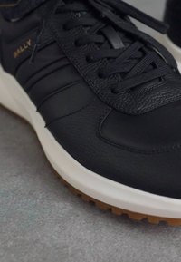 Zapatilla de cuero negro con punta redonda, superficie texturizada y cordones tonales. Presenta una entresuela blanca y una suela de goma color caramelo. Nombre de la marca "Bally" grabado.