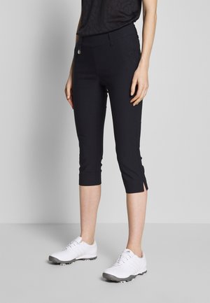 Femme portant un pantalon court noir avec des fentes latérales et des chaussures de sport blanches, debout devant un fond gris clair uni.
