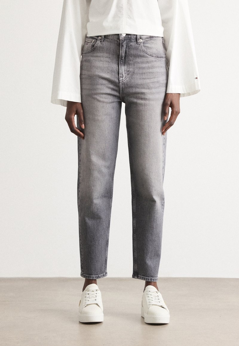 Personne portant un jean taille haute gris, un haut blanc à manches longues larges et des baskets blanches, debout sur un sol uni.