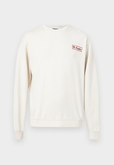 Μπεζ φούτερ με στρογγυλή λαιμόκοψη, μακριά μανίκια και μικρό ταμπελάκι με κόκκινο περίγραμμα στο αριστερό στήθος που γράφει "The Kooples".