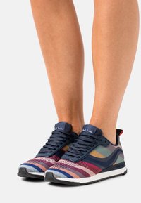 Sneakers con tomaia in rete a strisce multicolori. Presentano lacci blu marina, suola grigia e una linguetta sul tallone rossa a contrasto.