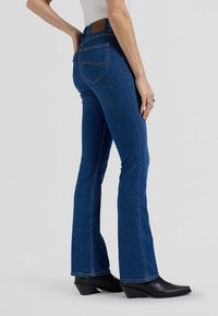 Žena u visokim plavim bootcut trapericama i crnim gležnjačama šiljastih vrhova, stoji s jednom rukom na kuku ispred jednobojne pozadine.