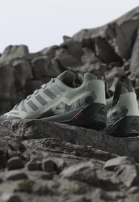Ανοιχτόγρια αθλητικά παπούτσια Adidas Terrex για τρέξιμο σε μονοπάτια με μαύρες σόλες, τοποθετημένα σε βραχώδες έδαφος σε εξωτερικό χώρο.
