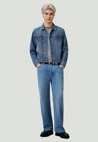 Young person with blonde hair wearing blue denim jacket, striped shirt, blue jeans, black belt, and black shoes, standing with hands in pockets.