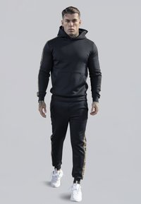 Black hoodie with a front pocket and gold accents along the sleeves, paired with matching black joggers featuring gold side stripes. White sneakers.