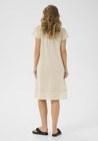 Robe en coton beige à manches courtes, longueur genoux, ornée d'un délicat détail brodé à l'ourlet. Assortie avec des sandales noires.