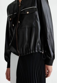 Giacca bomber nera in raso con chiusura a zip, due tasche sul petto e orlo elastico. Caratterizzata da dettagli in metallo dorato e profili bianchi.