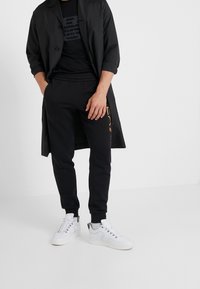 Musta kevyt takki napituksella, musta t-paita, jossa logo, mustat kapenevat verkkarit ja valkoiset lenkkarit hopeisilla yksityiskohdilla.