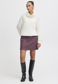 Pull en maille texturée avec col cheminée crème ; jupe violette à finition lisse, longueur au-dessus du genou ; bottes noires au genou avec talons carrés.