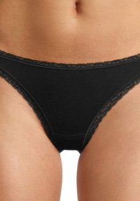 Slip nero in bikini realizzato in tessuto morbido, con un bordo in pizzo all'altezza della vita e delle aperture delle gambe, dal texture liscia.