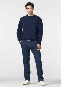 Sweatshirt bleu marine avec des poignets côtelés, associé à un jean bleu foncé et des baskets blanches. Logo rouge notable sur la poitrine du sweatshirt.