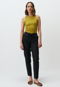 Olijfgroene geribbelde tanktop, zwarte linnen broek met trekkoord en bruine platte sandalen. Model staat tegen een effen witte achtergrond.