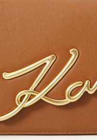 Brown leather handbag featuring a prominent gold metal logo in cursive script. Smooth texture with a subtle grain pattern.