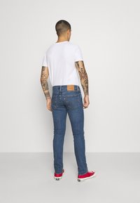 Vaqueros de mezclilla azul con un ajuste slim, que presentan un parche de cuero marrón en la parte trasera y zapatillas rojas. Camiseta blanca lisa llevada arriba.