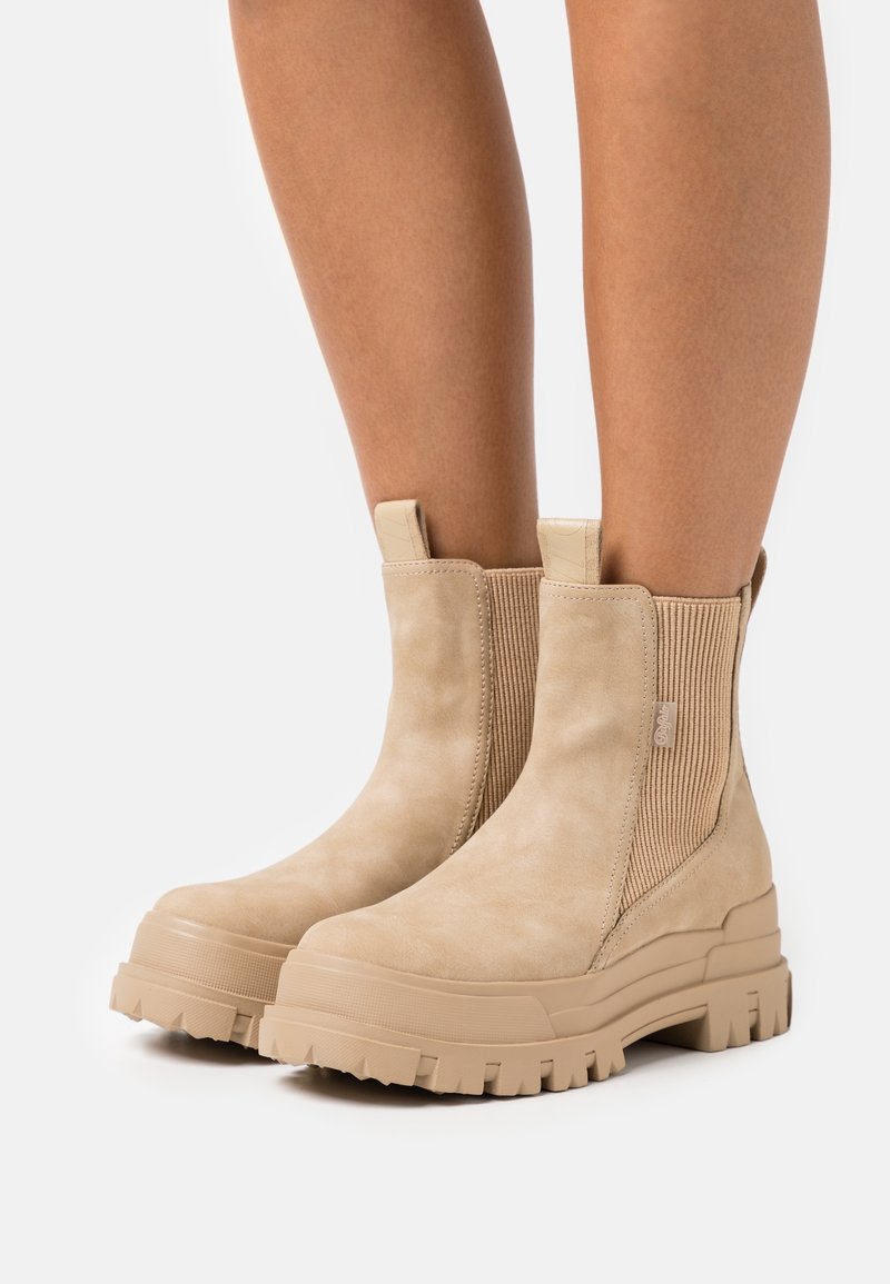 Bottines beiges en daim avec semelles épaisses à crampons et panneaux élastiques latéraux, portées sur des jambes nues devant un fond uni.