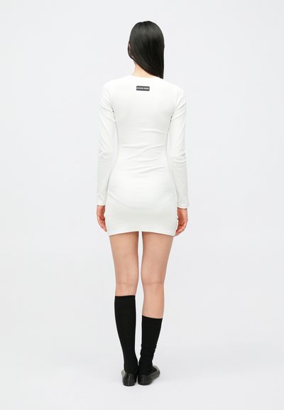 Biała sukienka z długim rękawem, dopasowanego kroju, z czarną metką z logo na szyi, w zestawie z czarnymi podkolanówkami i płaskimi butami.