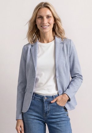 Femme aux cheveux blonds souriante, portant un blazer bleu clair, un t-shirt blanc et un jean bleu, une main dans la poche sur un fond uni.