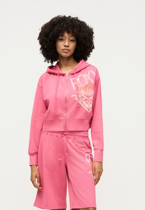 Femme aux cheveux bouclés portant un sweat à capuche zippé rose et un short assorti arrivant aux genoux avec texte graphique blanc et motifs.