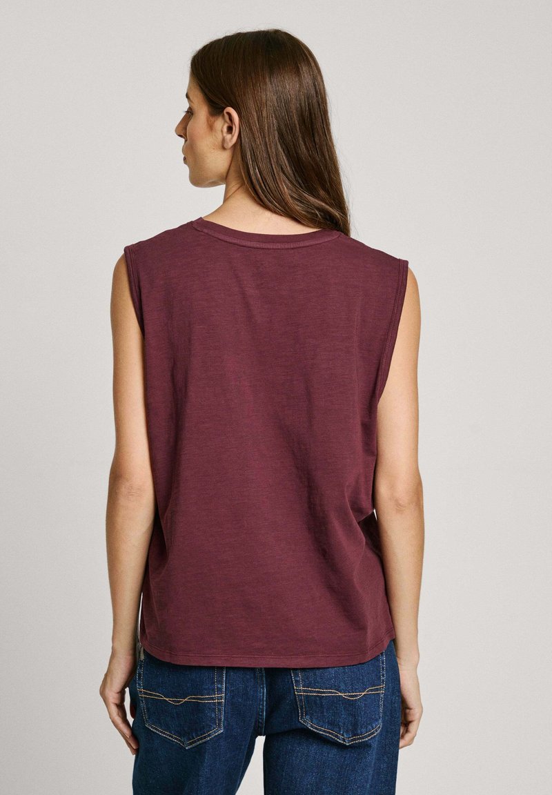 Bordeauxkleurige mouwloze top met een ronde hals, gemaakt van zachte stof. Beschikt over een relaxte pasvorm en een licht gebogen zoom, gecombineerd met donkerblauwe jeans.