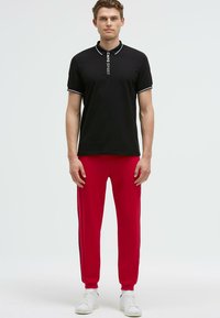 Camisa polo negra con ribete blanco y texto "CHMP SPORT", combinada con pantalones de chándal rojos con rayas laterales negras y zapatillas blancas.