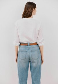 Camicia bianca a maniche lunghe con i polsini arrotolati, abbinata a jeans chiari a vita alta e una cintura marrone, vista da dietro.