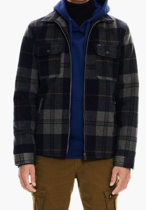 Mężczyzna ubrany w brązowe spodnie bojówki, niebieską bluzę z kapturem oraz rozpiętą czarno-szarą kurtkę w kratę z przednimi kieszeniami.